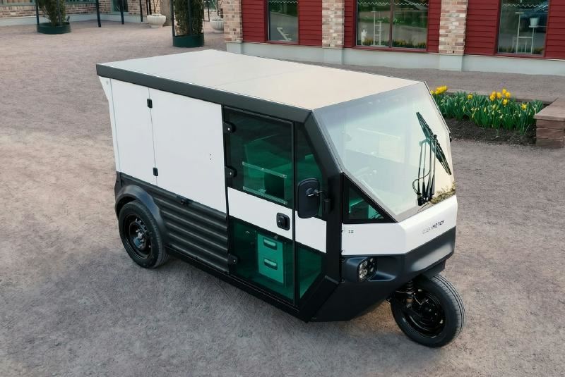 The image shows a compact, black electric delivery van with a green front compartment, parked in front of a red building with a green entrance.
AI-generated content may be incorrect.