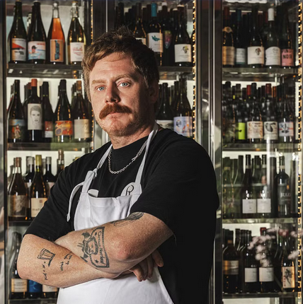 A person with a mustache and tattoos standing in front of a shelf of bottles
AI-generated content may be incorrect.
