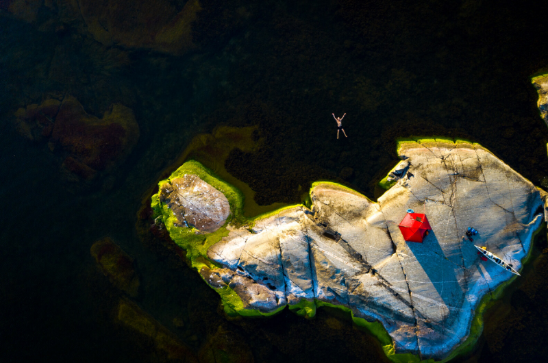 Luftaufnahme einer kleinen felsigen Insel in Schweden mit rotem Zelt, dunklem Wasser und einer schwimmenden Person. Das Bild vermittelt Ruhe, Abgeschiedenheit und naturnahe Reiseerlebnisse in den schwedischen Schären.